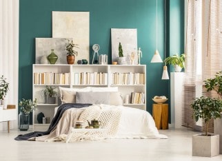 Bookcases with paintings on top standing against a dark green wall behind white bed in bright bedroom interior