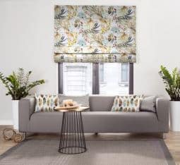 Salon w stylu urban jungle z wykorzystaniem motywów liści. Utrzymany w kolorystyce szarości i żółci. Charakteru nadaje wnętrzu roleta rzymska z kolekcji Urban Jungle oraz poduszki dekoracyjne z wzorami liści. Szary pokrowiec na sofę Klippan stanowi idealne tło dla aranżacji. Szyku dodaje tej aranżacji stolik ze złotym blatem.