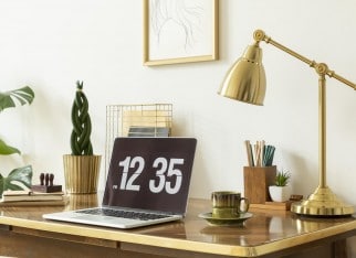 Gold lamp, laptop and plant on wooden desk in white freelancer's interior with poster. Real photo